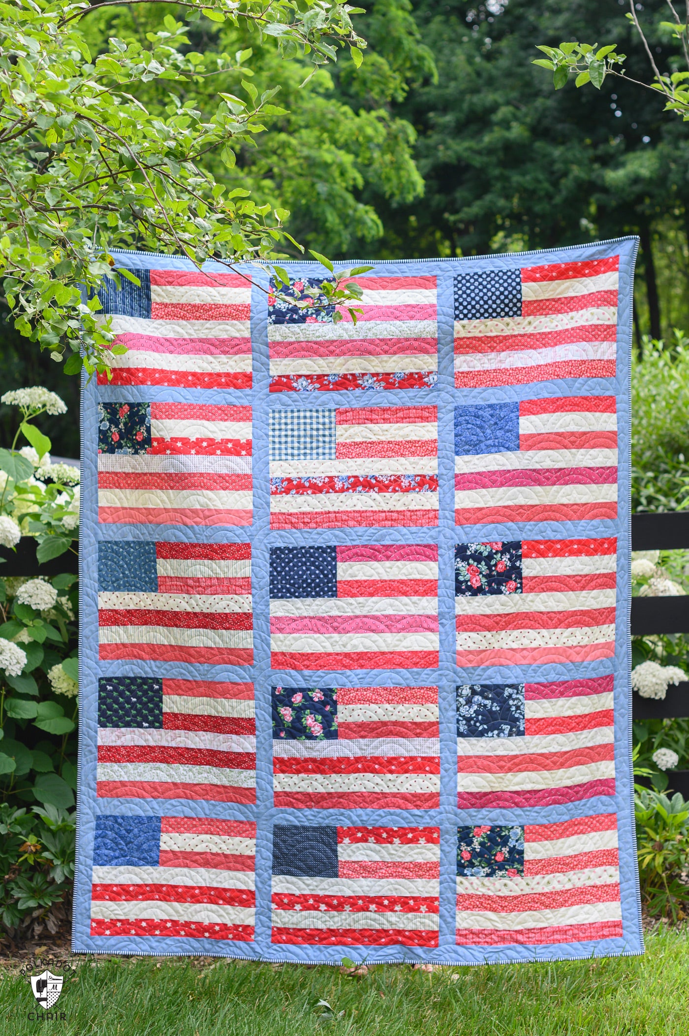 Quilt with American flag pattern displayed outdoors on a grassy area with greenery.