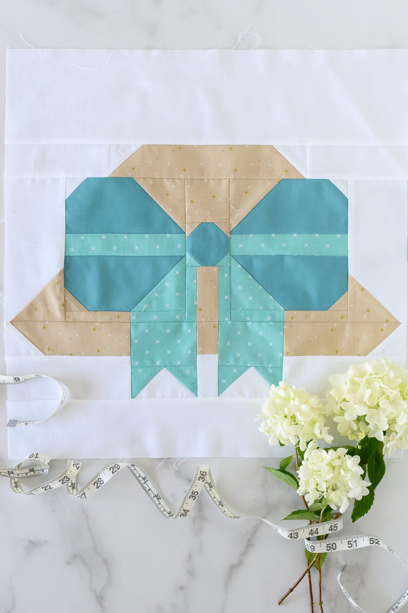Quilt block with a bow design on a white background with flowers and measuring tape.