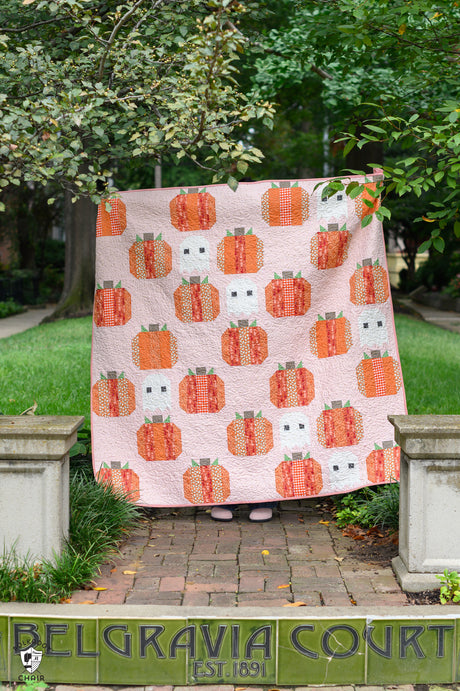 A pink quilted blanket featuring a repeating pattern of orange pumpkins and white ghosts, held up outdoors in a garden setting.