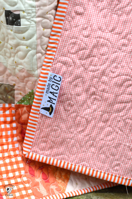 A close-up of a quilted pink and white gingham blanket with orange and white striped borders, featuring a 'made by MAGIC' brand tag.
